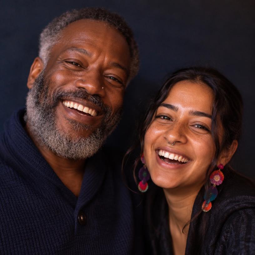 A joyful Black man in his early 60s with a gray beard and nose stud, laughing beside a freckled South Asian woman with a septum piercing
